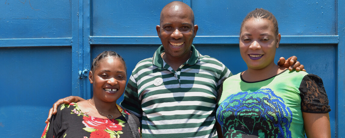 Two women and a man pose together smiling in front of a blue wall