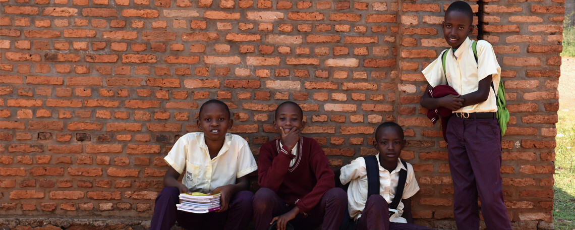 four school boys in uniform pose by a classroom wall with their books and bags