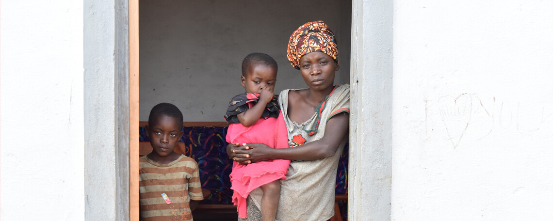 a woman holding a baby stand in the doorway of a new home with her son next to her