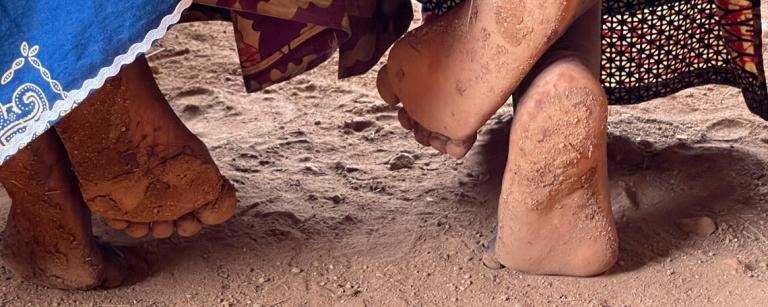 two ladies feet underneath a bench upon a dirt floor